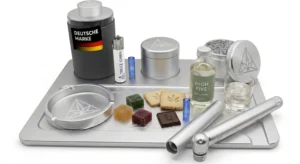 Komplettes Premium-Silber-Rolling-Tray-Set mit Cannabis-Zubehör, Grinder, Aschenbecher, THC-Gummies, THC-Keksen, THC-Schokolade, einer Flasche "HIGH FIVE" THC-Infusion und einem Glas Eiswasser vor weißem Hintergrund.